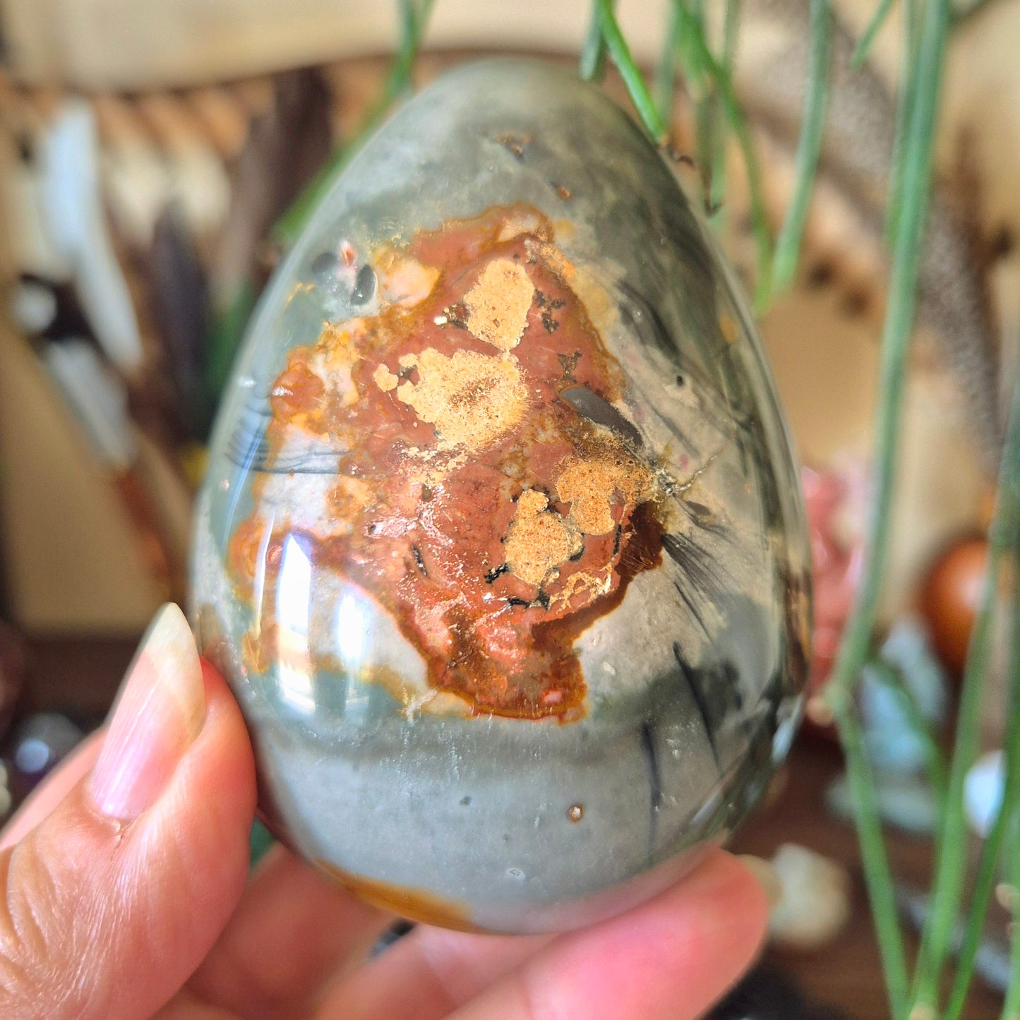 A close-up image of a Polychrome Jasper egg-shaped stone with a person's hand holding it.
