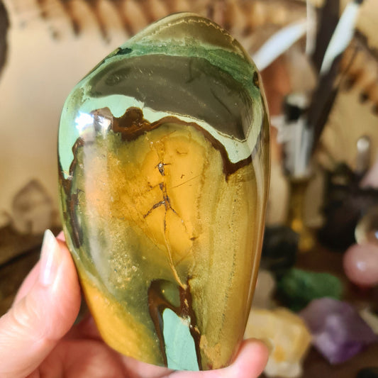 A polished freeform-shaped stone with a green body and brown/yellowish accents, held in a person's hand.