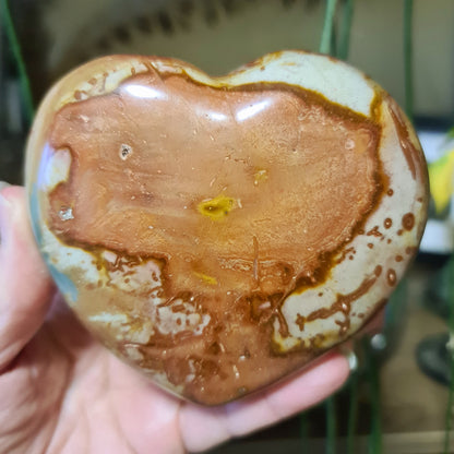 A hand holding a polychrome jasper heart-shaped stone with a brown and beige color pattern.