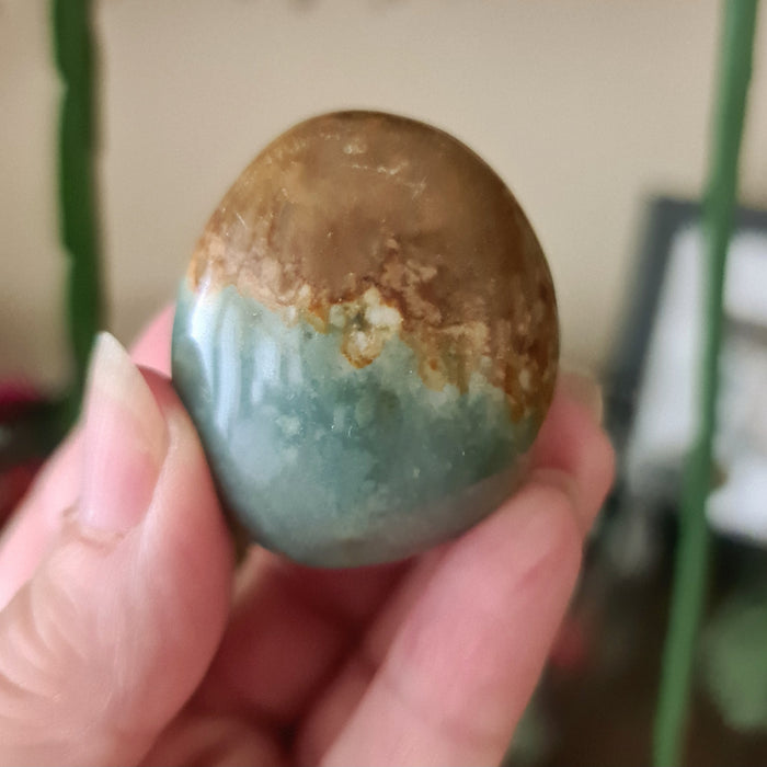 A polished palm-shaped stone with a blend of brown, green, and beige colors, held in a person's hand.