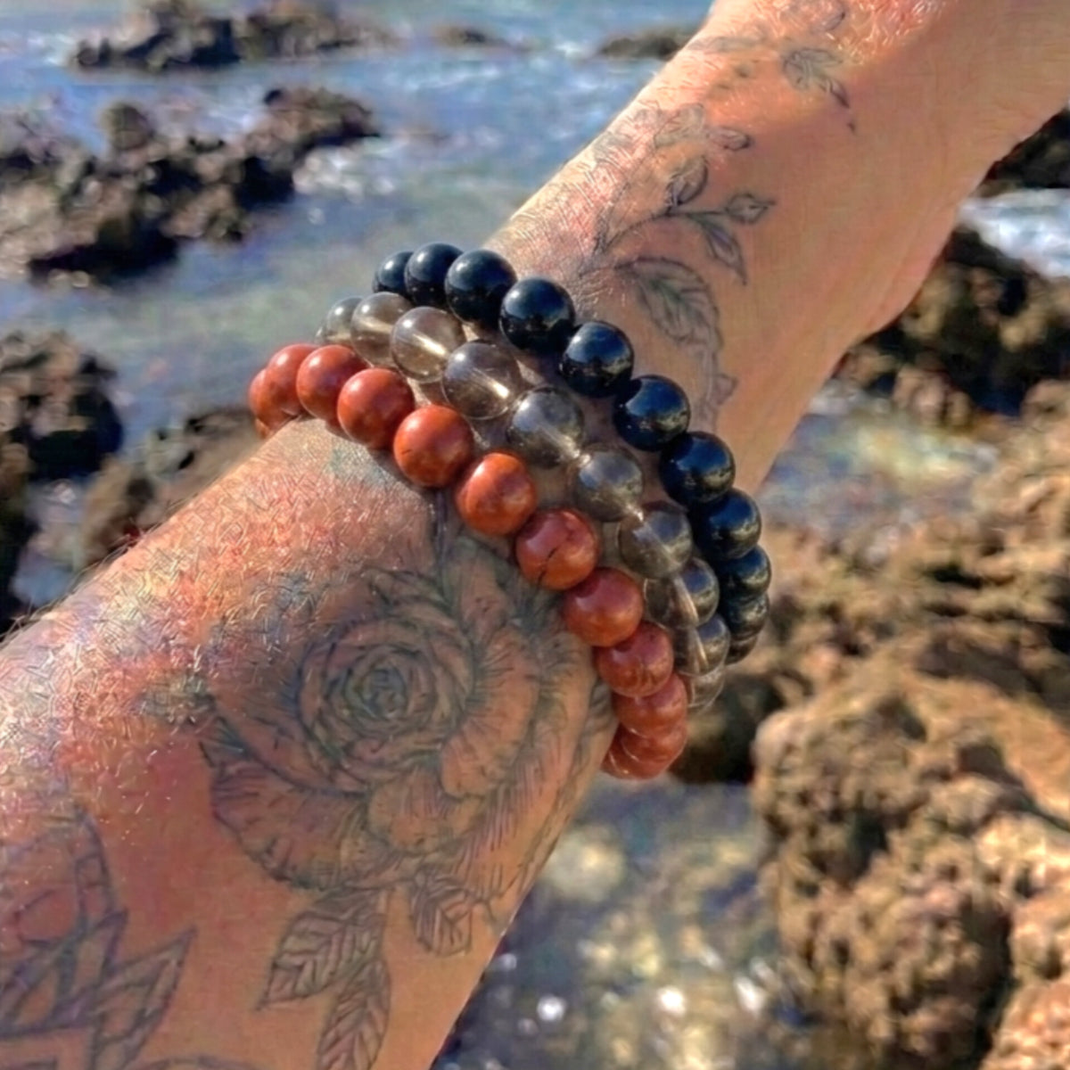 Three beaded bracelets on a tattooed wrist, featuring black obsidian, smoky quartz, and reddish-orange jasper beads, photographed outdoors against a rocky beach background.