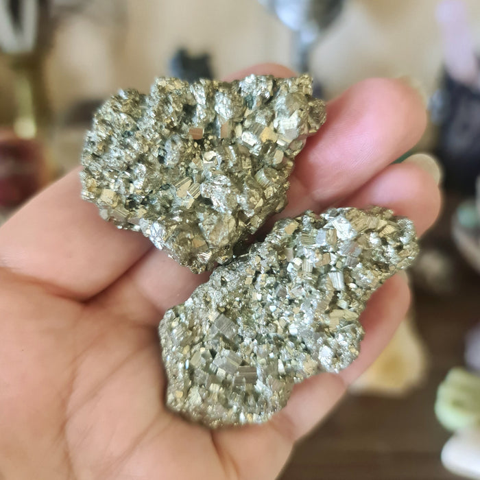 Rough pyrite crystal chunks held in a person's hand, with a blurred background suggesting indoor setting