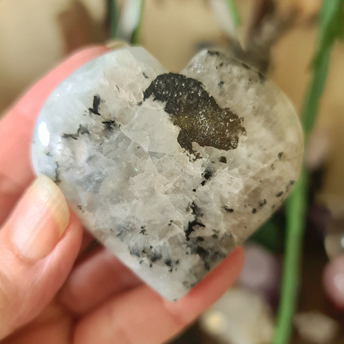 A heart-shaped rainbow moonstone with black and gray colors, held in a hand.
