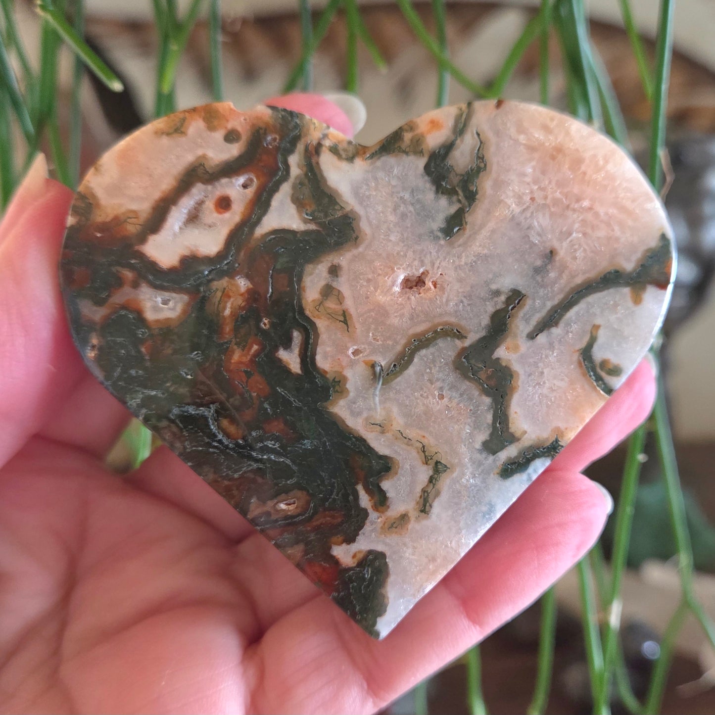 Heart-shaped stone with marbled pattern held in a hand, with green plants in the background