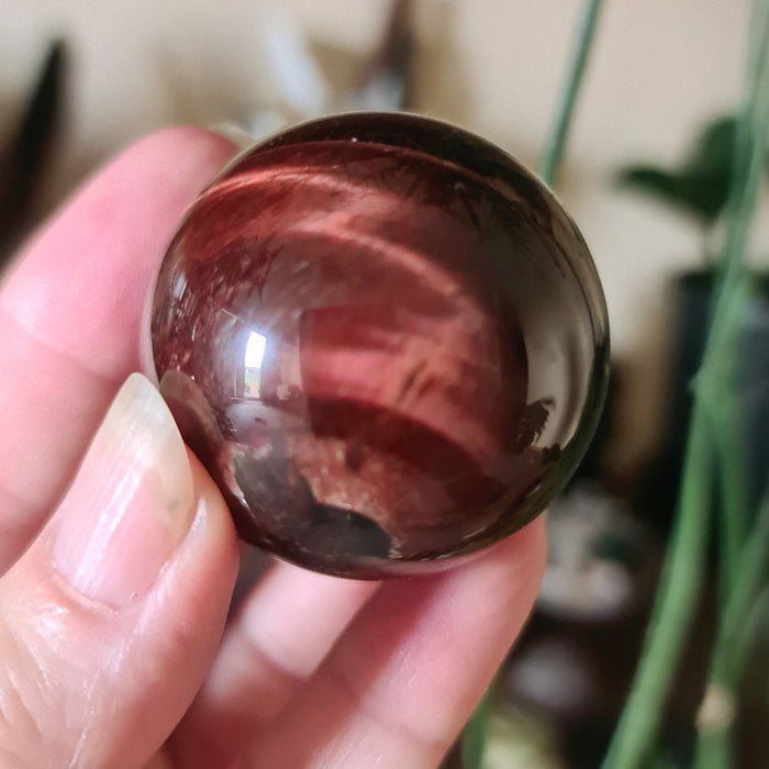 A hand holding a small, polished red tiger's eye stone with a smooth, glossy finish.