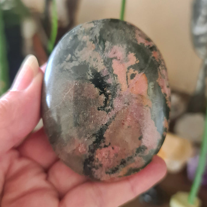 A polished Rhodonite Palm Stone with reddish and pinkish hues, held in a person's hand.