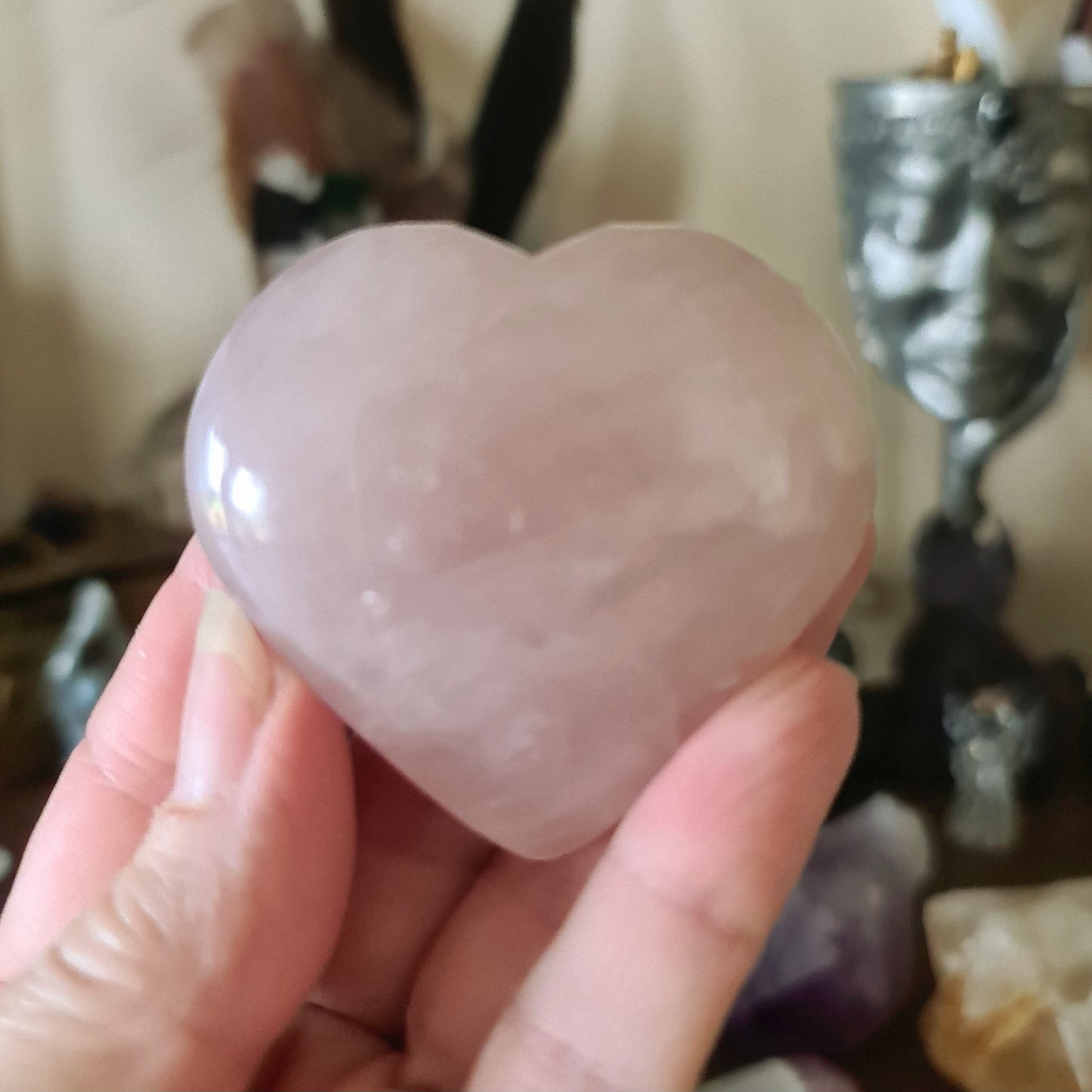 A large pink rose quartz heart-shaped crystal being held in a person's hand, with a blurred background featuring other crystals and a statue.