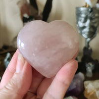 A large pink rose quartz heart-shaped crystal being held in a person's hand, with a blurred background featuring other crystals and a statue.