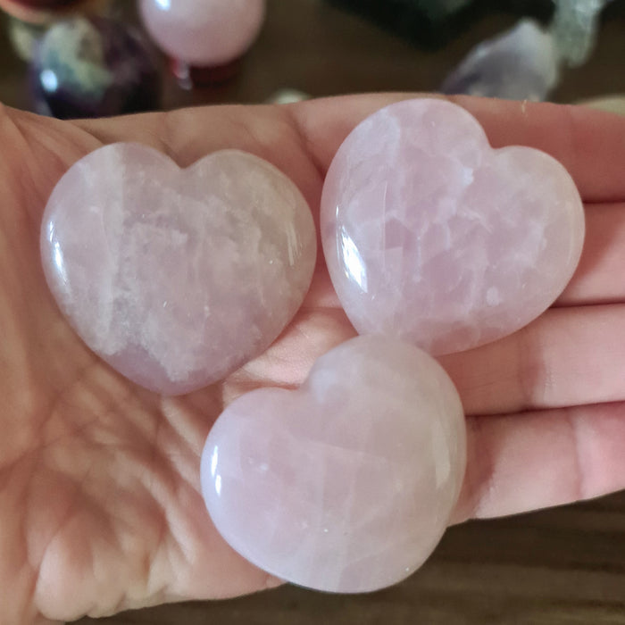A hand holding three polished pink rose quartz crystals shaped like hearts, with a blurred background.
