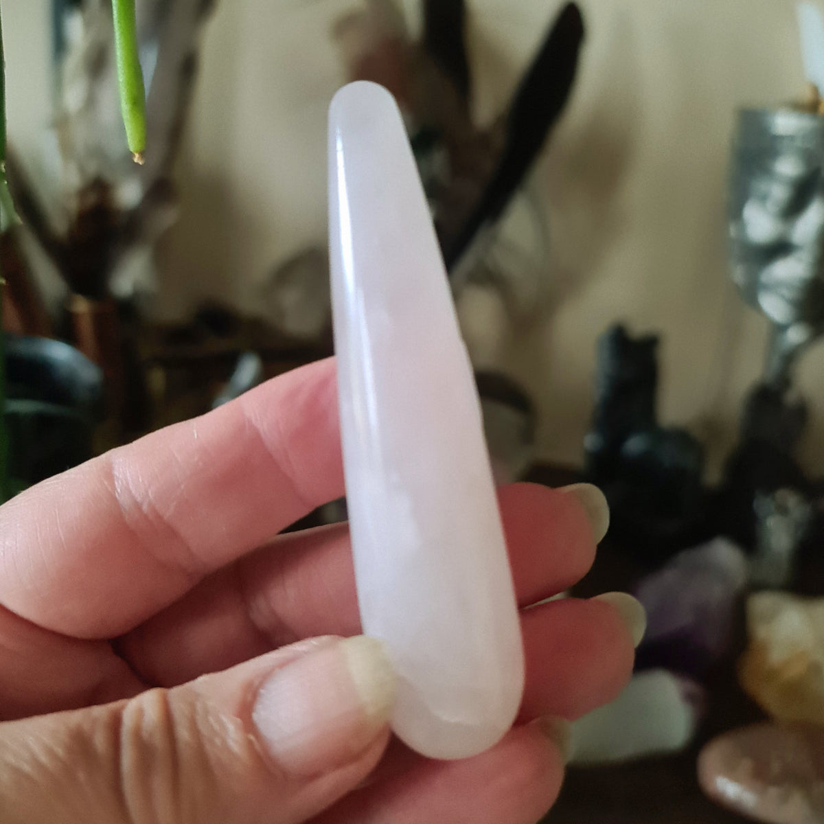 A hand holding a pink-colored Rose Quartz massage wand, with a blurred background featuring various crystal stones and decorative items.