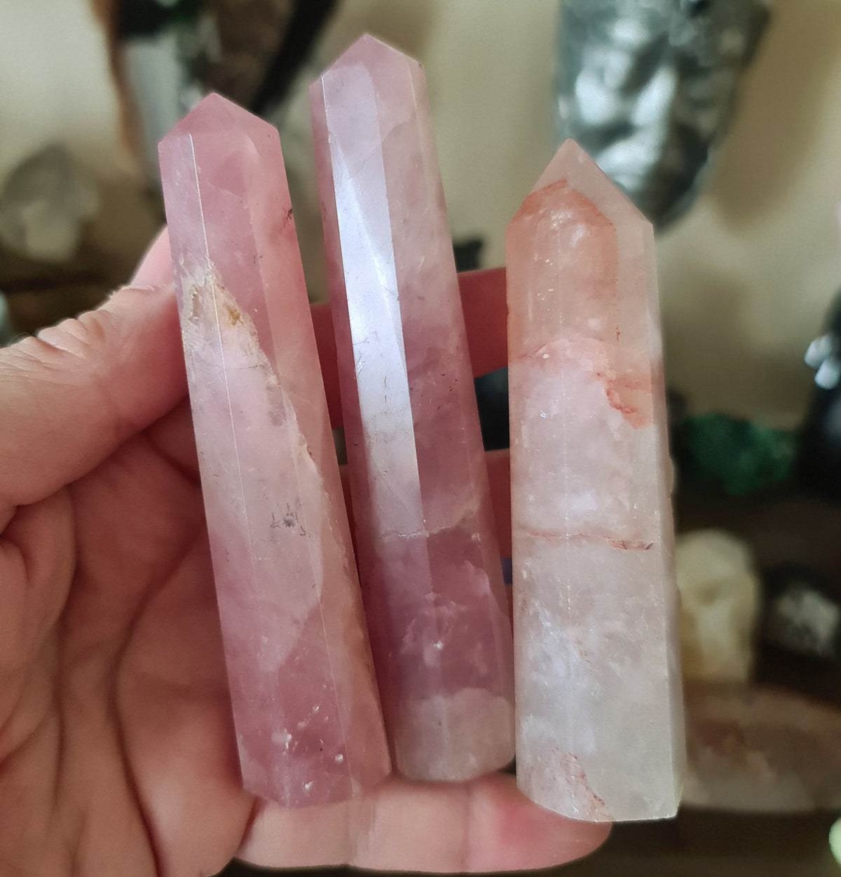 A hand holding a polished pink rose quartz crystal point, with a few drops of water visible on the surface.