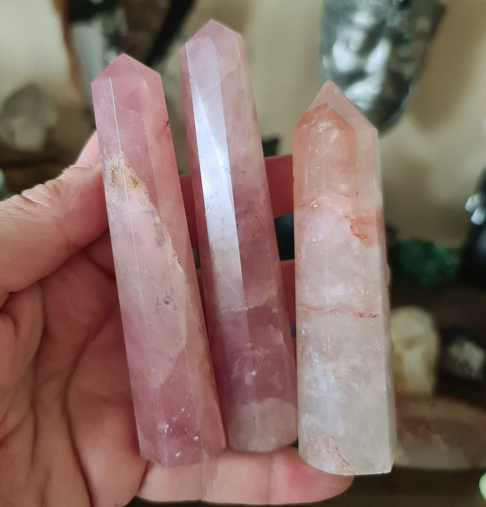 A hand holding a polished pink rose quartz crystal point, with a few drops of water visible on the surface.