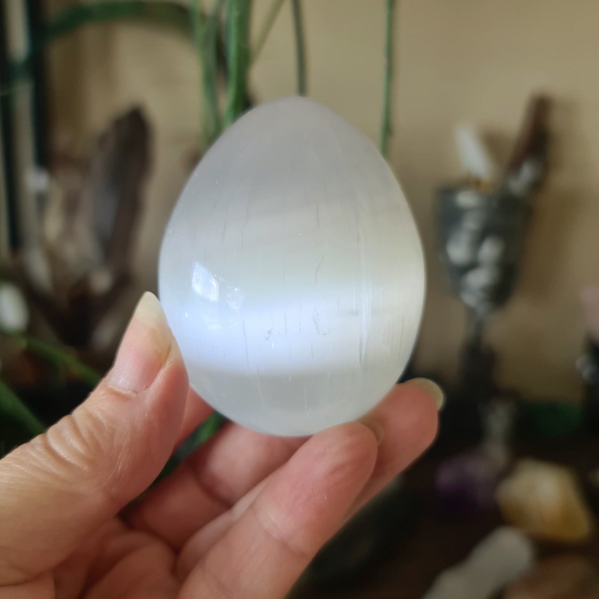 A white, egg-shaped crystal stone held in a person's hand, with a blurred background suggesting it might be a selenite egg.