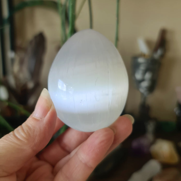 A white, egg-shaped crystal stone held in a person's hand, with a blurred background suggesting it might be a selenite egg.