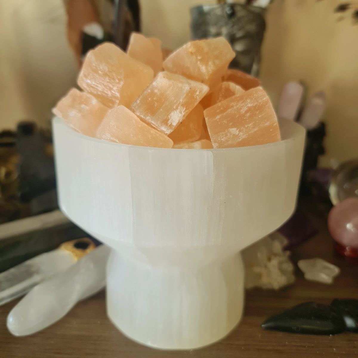 A white selenite crystal bowl lamp with peach selenite cubes, placed on a wooden surface, with various crystals and items in the background.