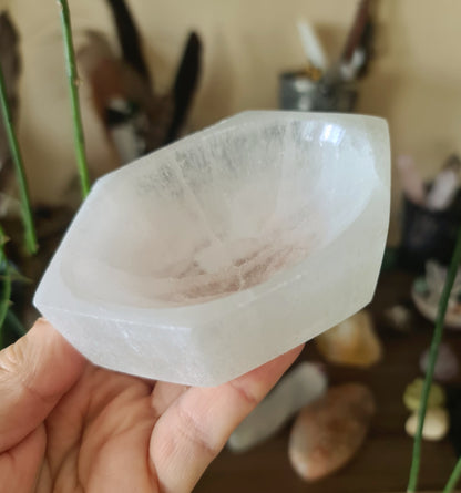 A clear, flat, bowl-shaped selenite crystal holder, held in a person's hand, with a blurred background suggesting a home or personal item.