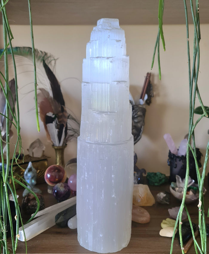 A large, clear selenite crystal lamp with a light bulb, handcrafted and ethically sourced, displayed on a table with various crystals and decorative items in the background.