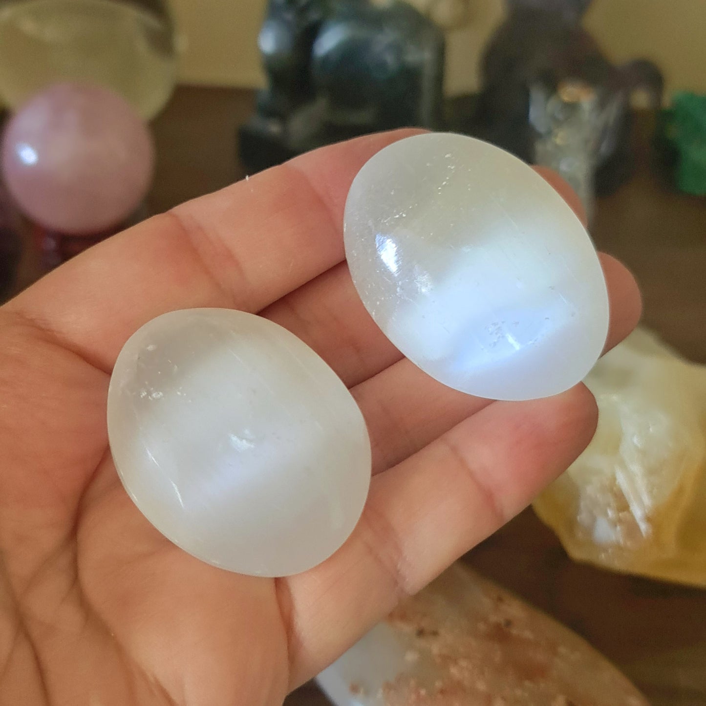 A pair of white, oval-shaped selenite crystal figures held in a person's hand, with various other crystals and spiritual items in the background.