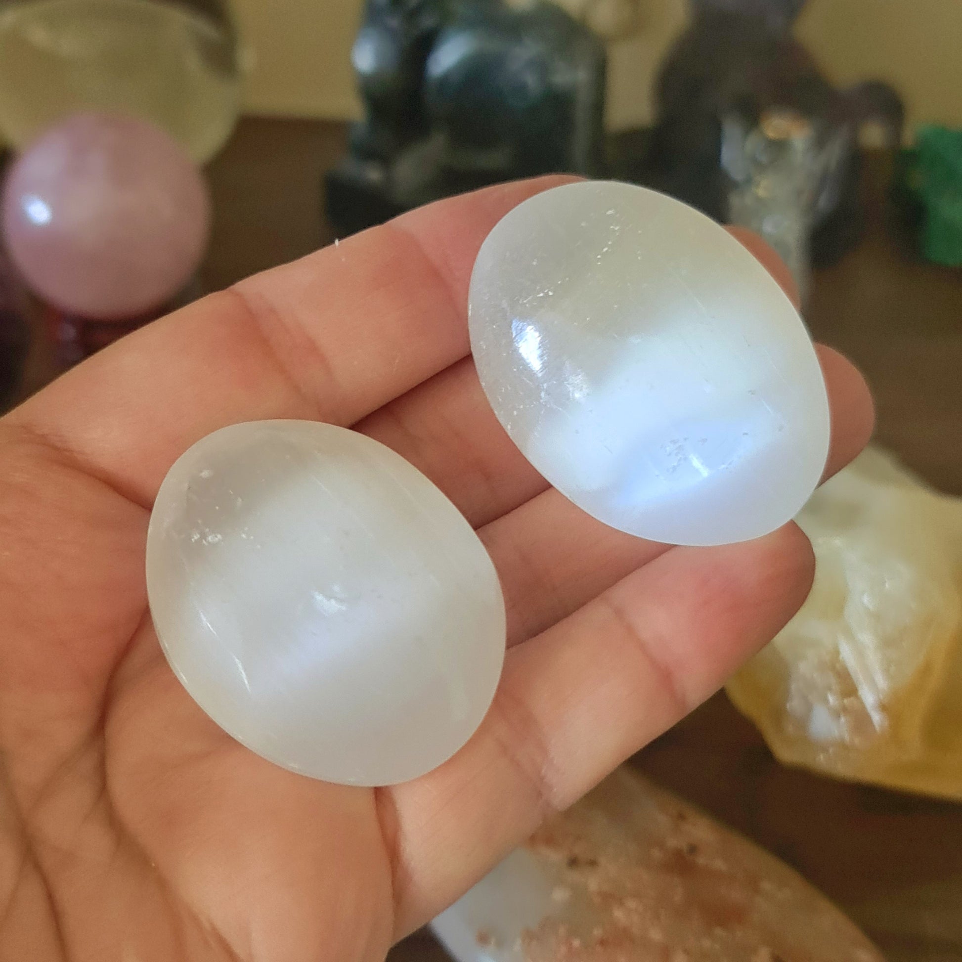 A pair of white, oval-shaped selenite crystal figures held in a person's hand, with various other crystals and spiritual items in the background.