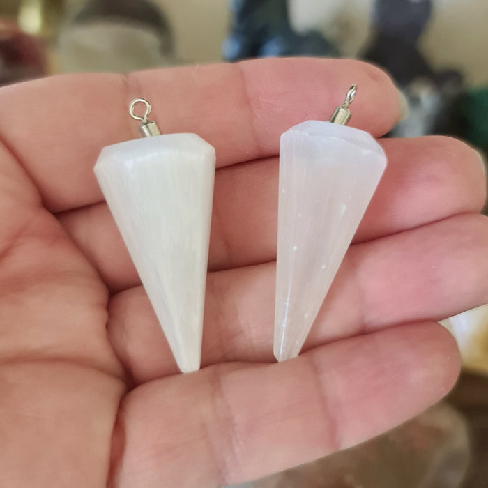 Two white crystal pendulums held in a person's hand, with metal loops attached to their top ends.