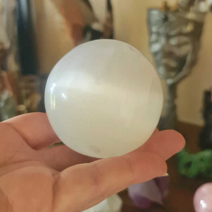 A medium-sized, translucent white selenite sphere being held in a person's hand, with blurred objects in the background.