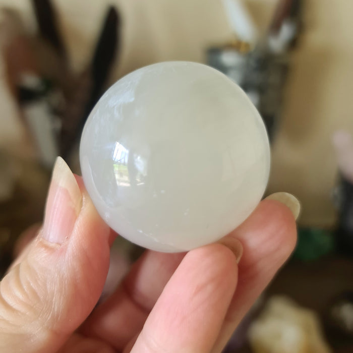 A small white selenite sphere held in a person's hand, with a blurred background suggesting a indoor setting