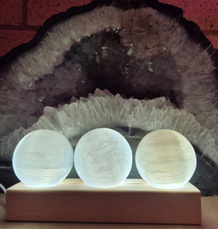 Three clear selenite spheres on a wooden trio LED base, with the base illuminated by the LED lights.