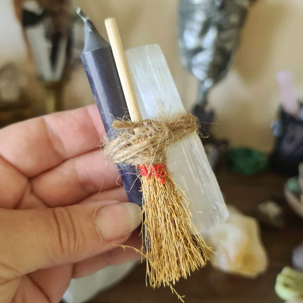 A three-piece Selenite Sweep Set, consisting of a broom, a black candle, and a clear crystal, held in a person's hand.