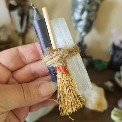 A three-piece Selenite Sweep Set, consisting of a broom, a black candle, and a clear crystal, held in a person's hand.