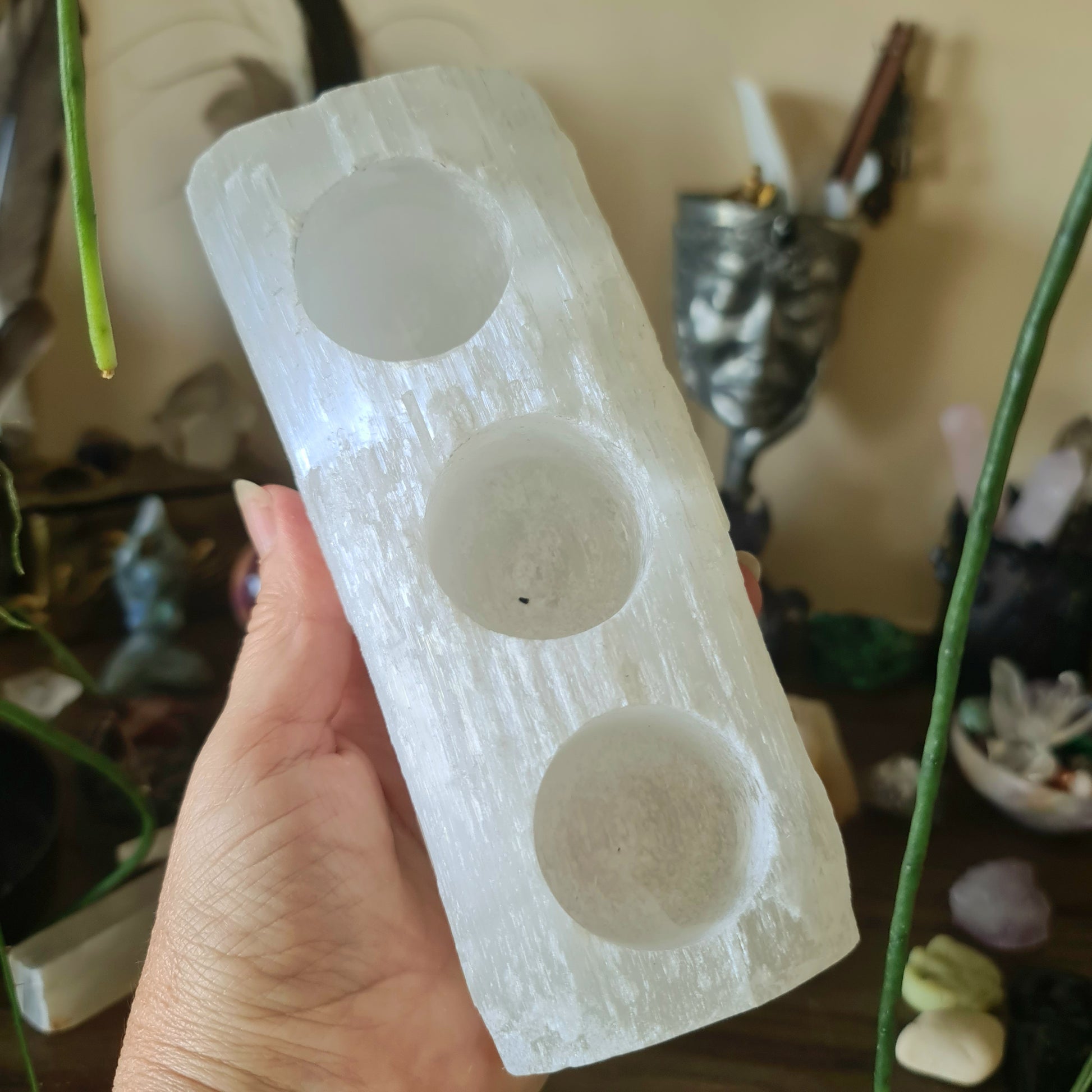 A hand holding a raw white selenite crystal tealight holder with three circular indentations for candles.