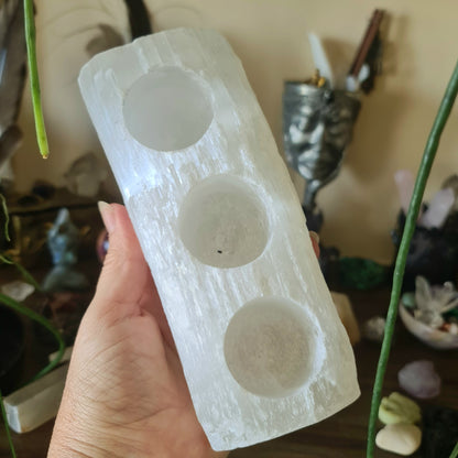 A hand holding a raw white selenite crystal tealight holder with three circular indentations for candles.