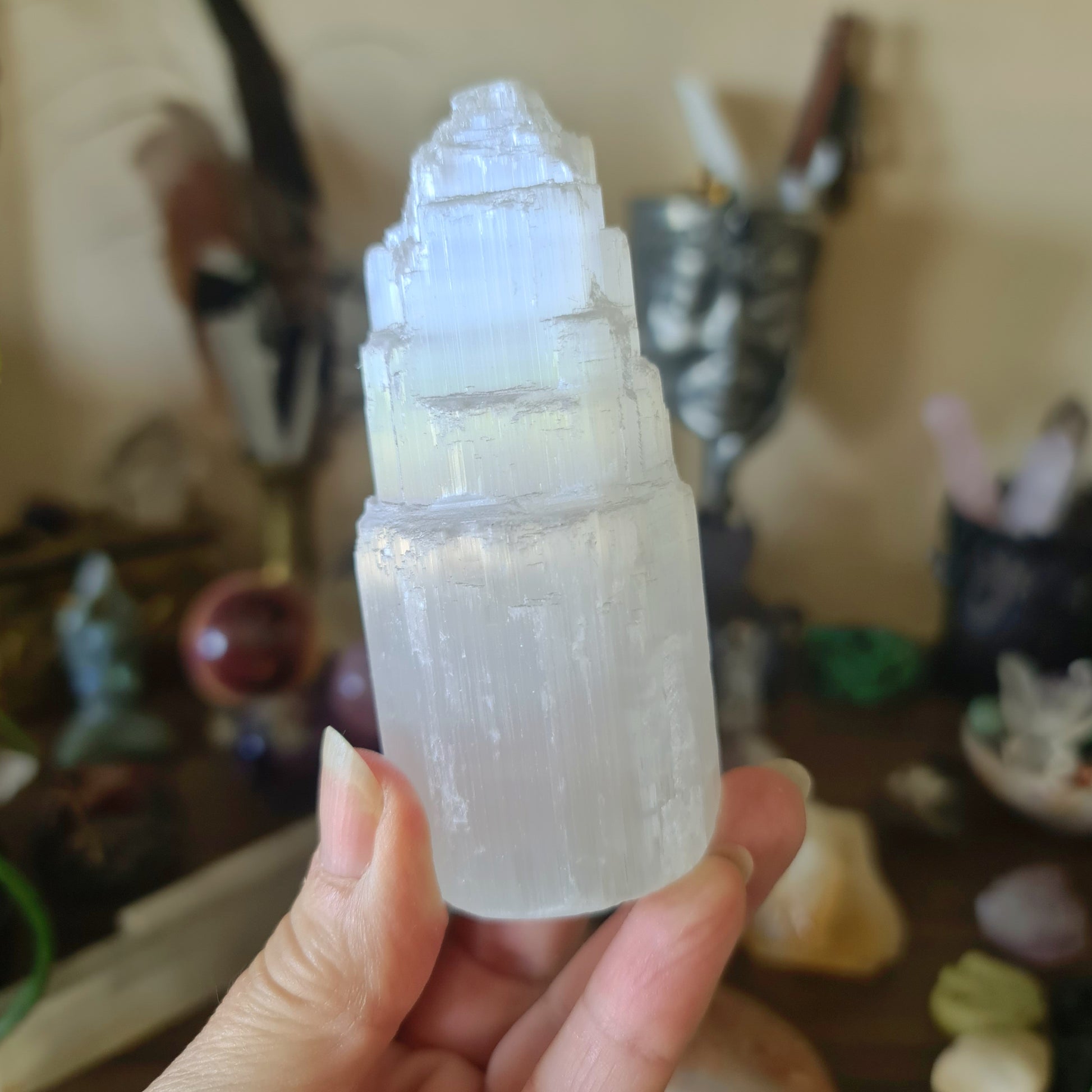 A small, clear selenite crystal tower held in a person's hand, with various other crystals and items in the background on a table.