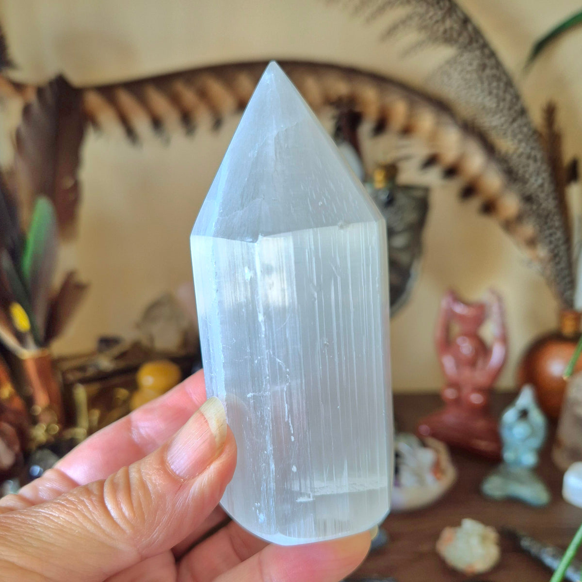 A hand holding a translucent selenite crystal tower, with a blurred background containing various decorative items.
