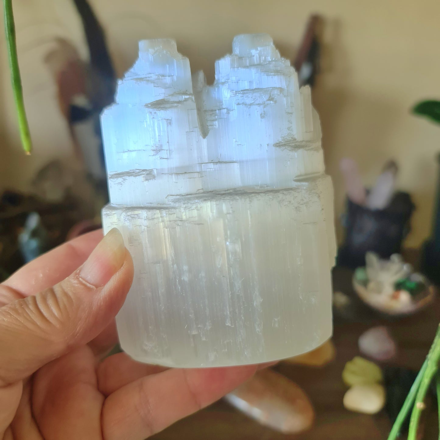 A clear, twin-tower selenite crystal figure held in a person's hand, with a blurred background suggesting a home or meditation setting.