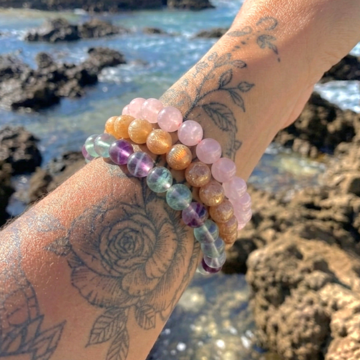 A close-up of a tattooed wrist wearing multiple beaded bracelets outdoors by the rocky ocean shore. The bracelets include translucent pink, peach, lavender, green, blue, and metallic gold beads.
