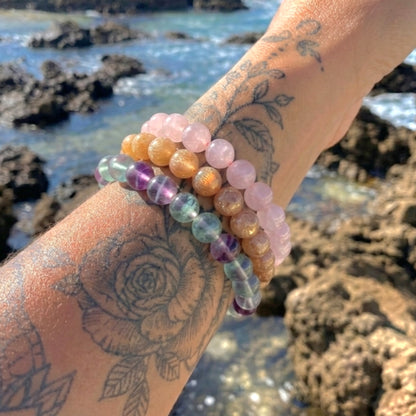 A close-up of a tattooed wrist wearing multiple beaded bracelets outdoors by the rocky ocean shore. The bracelets include translucent pink, peach, lavender, green, blue, and metallic gold beads.