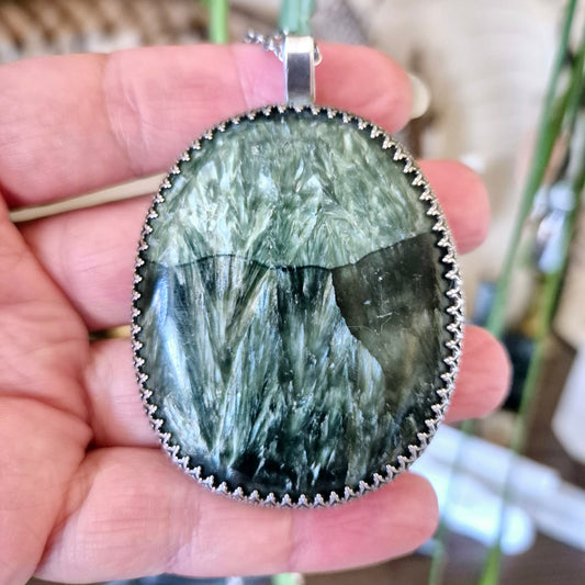 A hand holding a silver pendant with a green seraphinite stone.