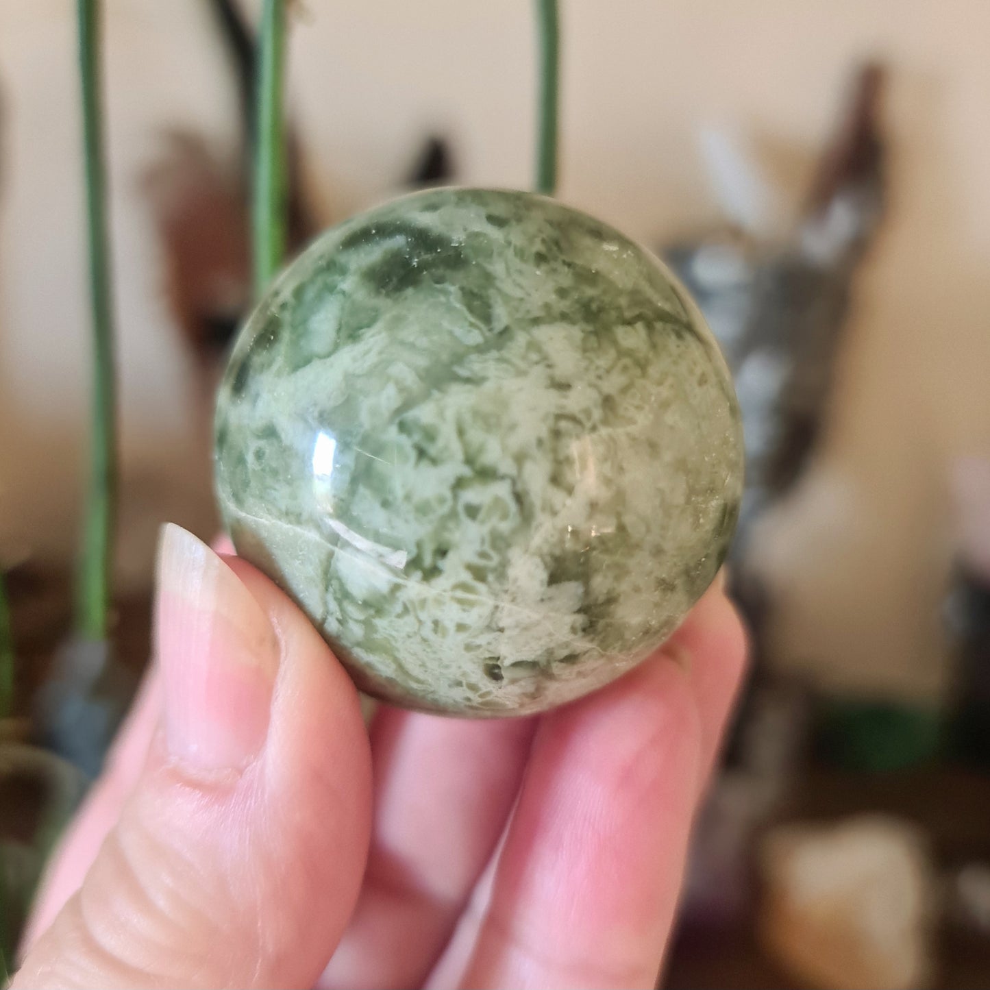 A polished green serpentine sphere held in a person's hand, with a blurred background suggesting indoor setting