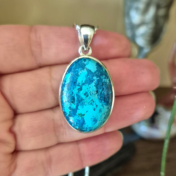 A sterling silver pendant featuring an oval-shaped blue Shattuckite stone with a metallic and glass-like appearance, held in a person's hand.