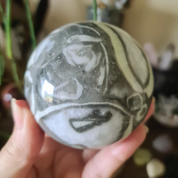 A spherical Shell Jasper stone with a design that resembles a stylized face, held in a person's hand.
