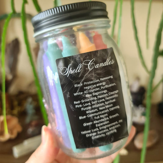 A clear jar containing multiple colored candles, with a label that lists the candle colors and their meanings: black, white, red, pink, purple, blue, green, and yellow.