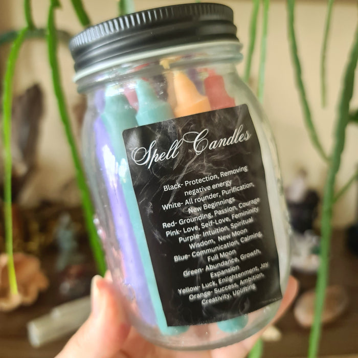 A clear jar containing multiple colored candles, with a label that lists the candle colors and their meanings: black, white, red, pink, purple, blue, green, and yellow.