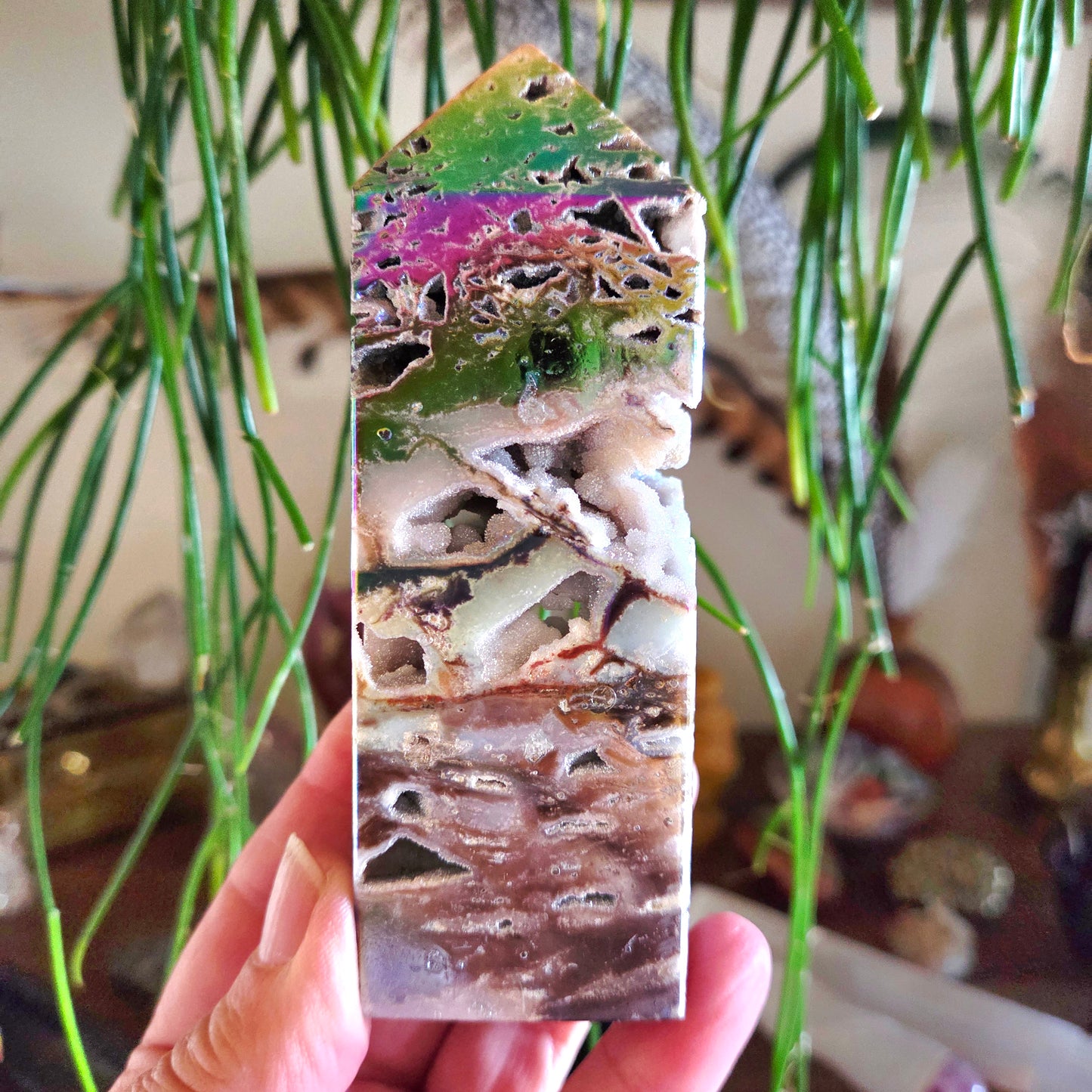 A hand holding a multicolored mineral crystal with a tower shape, displaying hues of green, pink, and brown.