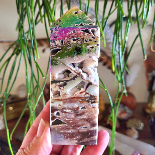 A hand holding a multicolored mineral crystal with a tower shape, displaying hues of green, pink, and brown.
