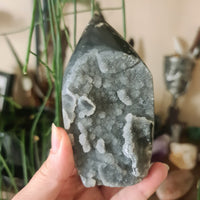 A hand holding a polished Sphalerite Free Form crystal, with a blurred background suggesting a indoor setting, possibly a store or home environment.