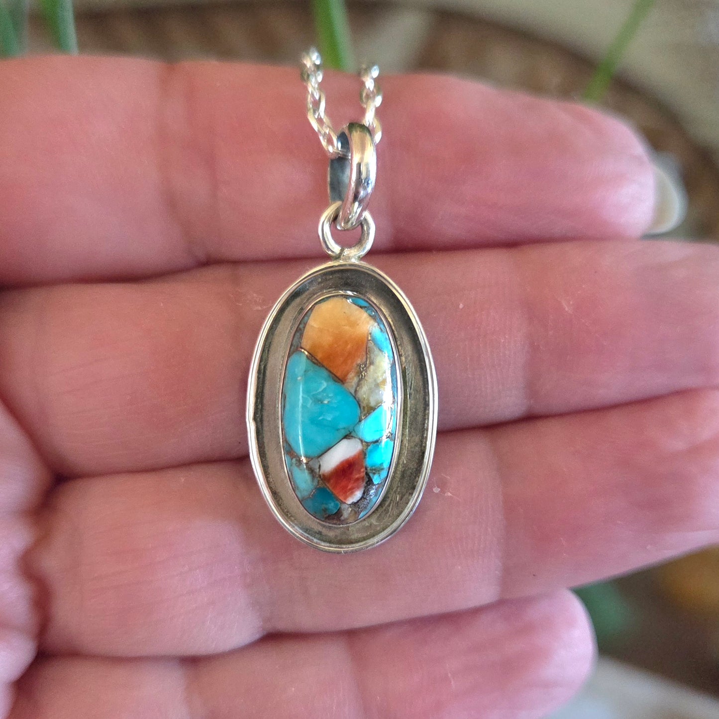 Turquoise and coral pendant on a silver chain held in a hand.