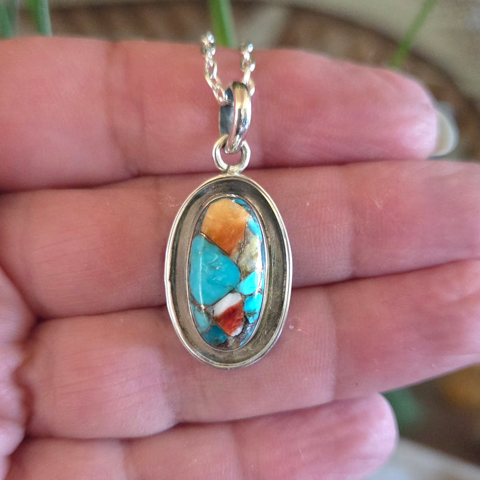 Turquoise and coral pendant on a silver chain held in a hand.