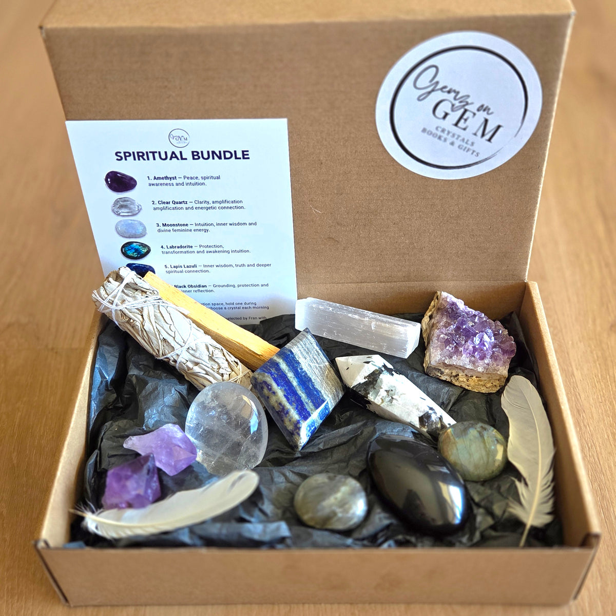An open cardboard gift box labeled containing a spiritual bundle with crystals, a smudge stick, a feather, and a printed 'SPIRITUAL BUNDLE' card on black tissue paper.