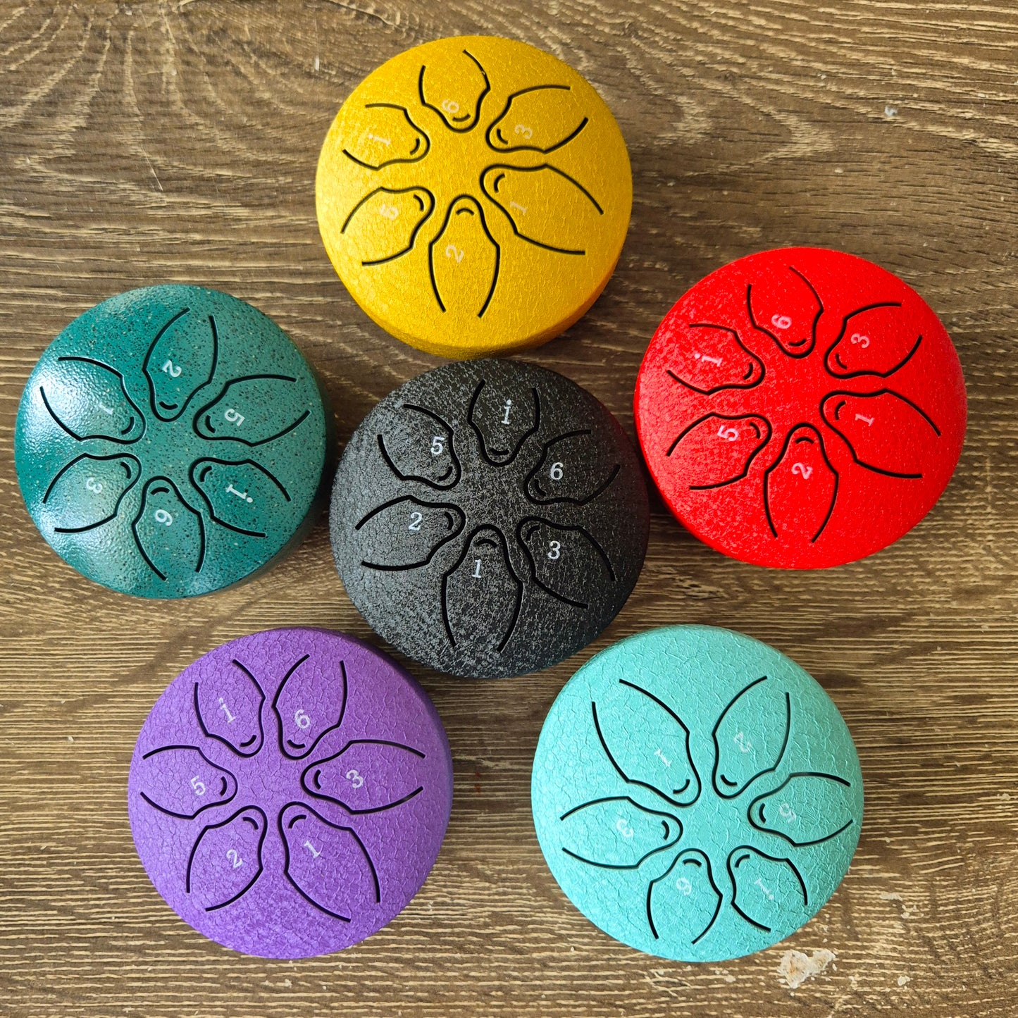 A set of five steel tongue drums in various colors placed on a wooden surface, with each drum having a floral pattern and a number indicating pitch.