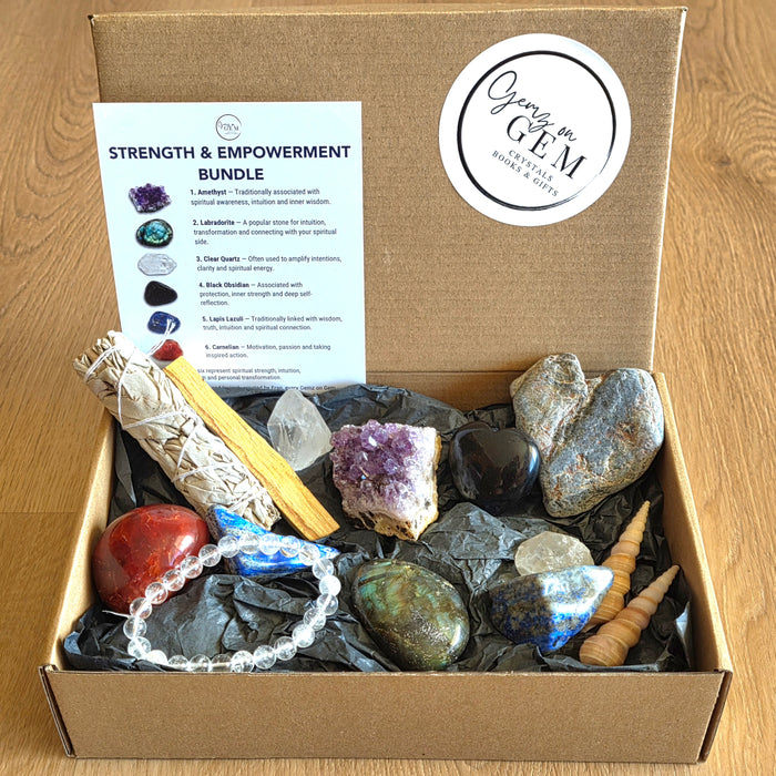 A 'Strength & Empowerment Bundle' GEM box set displayed in an open cardboard box on a wooden table. The set includes a white sage bundle, a red jasper stone, a blue tourmaline bead strand, a purple amethyst geode, a black obsidian stone, a blue lapis lazuli crystal, a gray agate stone, a white quartz crystal, a spiral shell, and an informational card detailing the contents.
